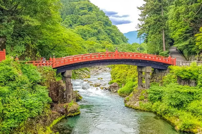 A vivid red Shinkyo Bridge arching over the clear waters of the Daiya River, surrounded by lush green forest in Nikko, Japan.